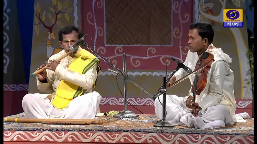 Two musicians sit cross-legged, one playing a flute, the other a violin. They're on a stage, performing for DD Bangla, the channel's logo visible in the corner.
Two musicians sit cross-legged, one playing a flute, the other a violin. They're on a stage, performing for DD Bangla, the channel's logo visible in the corner.
