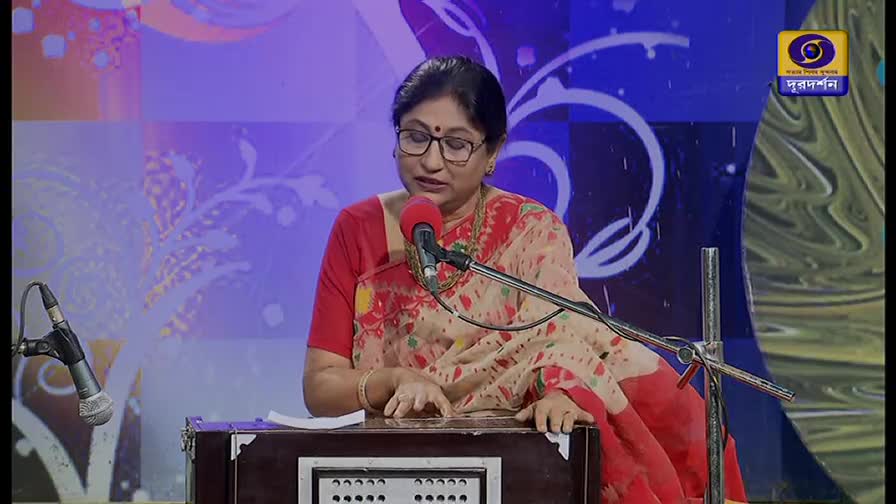A woman in a red and cream sari sits before a harmonium, her fingers moving over the keys. She's singing into a microphone, likely on DD Bangla, with the Doordarshan logo visible in the corner.
A woman in a red and cream sari sits before a harmonium, her fingers moving over the keys. She's singing into a microphone, likely on DD Bangla, with the Doordarshan logo visible in the corner.