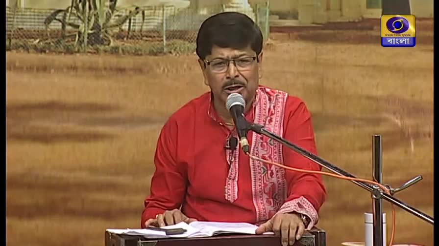 A man in a red shirt and glasses sings into a microphone, his hands resting on a small instrument. The DD Bangla logo is visible in the corner.
A man in a red shirt and glasses sings into a microphone, his hands resting on a small instrument. The DD Bangla logo is visible in the corner.