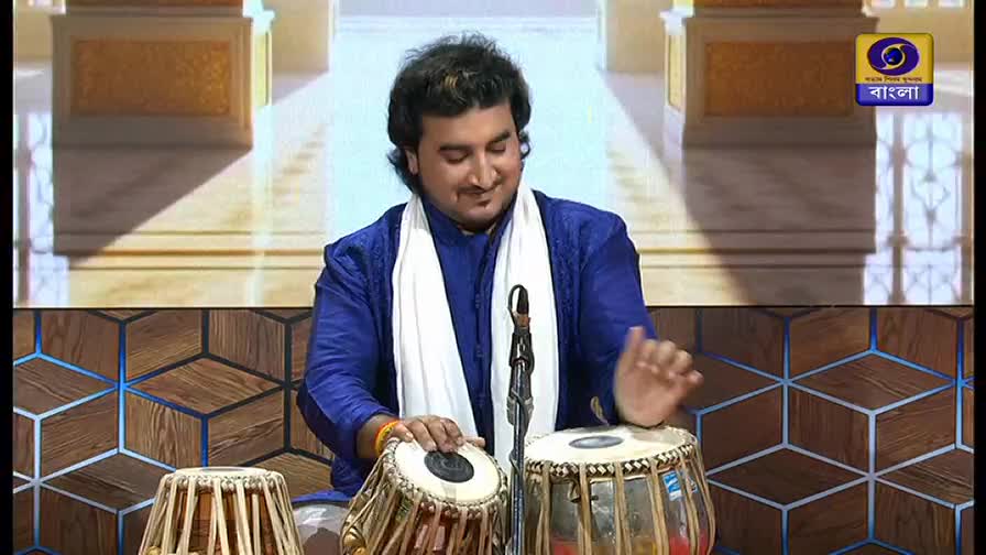 The musician, wearing a blue kurta and white scarf, smiles as his hands dance across the tabla drums. The scene is lit for a performance on DD Bangla, the logo visible in the corner.
The musician, wearing a blue kurta and white scarf, smiles as his hands dance across the tabla drums. The scene is lit for a performance on DD Bangla, the logo visible in the corner.