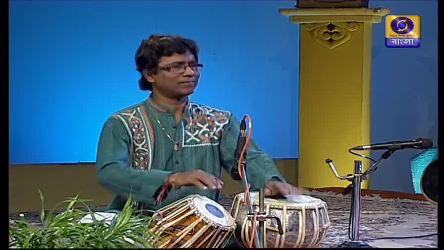 The musician's hands are a blur as they strike the tabla drums. He's on a DD Bangla set, with the channel's logo visible in the corner.
The musician's hands are a blur as they strike the tabla drums. He's on a DD Bangla set, with the channel's logo visible in the corner.