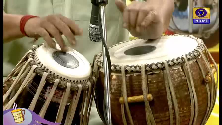 A musician's hands dance over the tabla drums, fingers striking the white surfaces. The DD Bangla logo is visible in the corner, suggesting a broadcast from India.
A musician's hands dance over the tabla drums, fingers striking the white surfaces. The DD Bangla logo is visible in the corner, suggesting a broadcast from India.