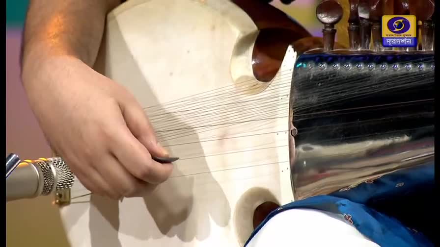 A musician's hand holds a small pick, strumming the instrument's strings. The camera angle is close, and the DD Bangla logo is visible in the background.
A musician's hand holds a small pick, strumming the instrument's strings. The camera angle is close, and the DD Bangla logo is visible in the background.