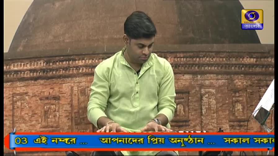 A man in a light green shirt is focused on playing a keyboard, his fingers moving across the keys. Behind him, the backdrop is a weathered, brown brick structure, and the DD Bangla logo is visible in the corner.
A man in a light green shirt is focused on playing a keyboard, his fingers moving across the keys. Behind him, the backdrop is a weathered, brown brick structure, and the DD Bangla logo is visible in the corner.