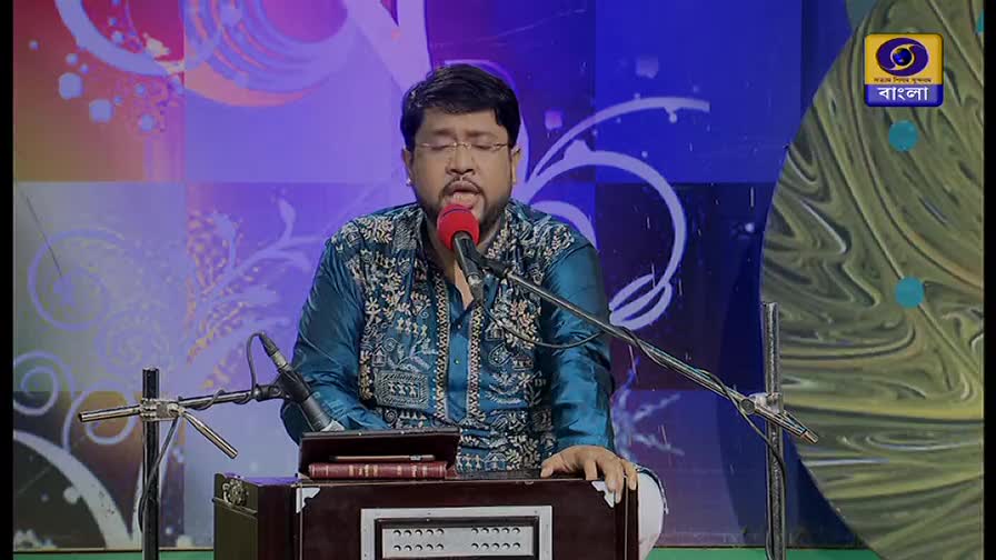 A man with glasses sings into a red microphone, his hands resting on a harmonium. The DD Bangla logo is visible in the corner, and the background is a swirl of colors.
A man with glasses sings into a red microphone, his hands resting on a harmonium. The DD Bangla logo is visible in the corner, and the background is a swirl of colors.