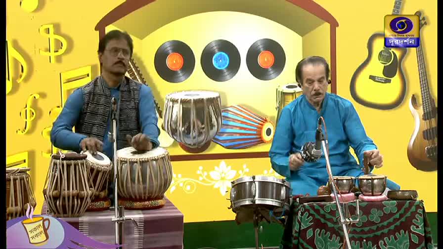 Two men, seated on a stage, are playing percussion instruments. The man on the left is striking the tabla, while the other man taps a smaller set of drums, all under the logo of DD Bangla.
Two men, seated on a stage, are playing percussion instruments. The man on the left is striking the tabla, while the other man taps a smaller set of drums, all under the logo of DD Bangla.