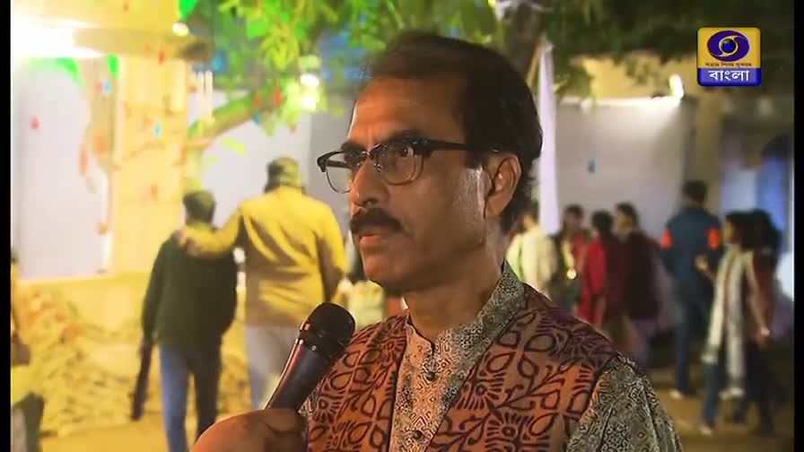 A man in glasses holds a microphone, speaking into it, with the DD Bangla logo visible in the corner. Behind him, people mill about in a brightly lit outdoor setting.
A man in glasses holds a microphone, speaking into it, with the DD Bangla logo visible in the corner. Behind him, people mill about in a brightly lit outdoor setting.