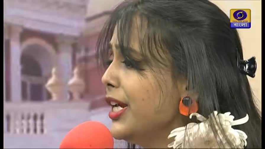 A woman with dark hair and bright red lipstick speaks into a microphone, her face animated. The DD Bangla logo sits in the corner, and a blurred backdrop suggests an outdoor setting.
A woman with dark hair and bright red lipstick speaks into a microphone, her face animated. The DD Bangla logo sits in the corner, and a blurred backdrop suggests an outdoor setting.