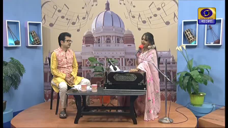 A woman in a pink sari sings into a microphone, playing a harmonium. Beside her, a man in a yellow kurta sits and watches, the backdrop a painted scene with musical notes and a DD Bangla logo.
A woman in a pink sari sings into a microphone, playing a harmonium. Beside her, a man in a yellow kurta sits and watches, the backdrop a painted scene with musical notes and a DD Bangla logo.