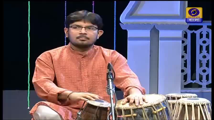 A musician in an orange kurta sits cross-legged, his hands poised over a pair of tablas. The DD Bangla logo is visible in the corner, indicating a broadcast from India.
A musician in an orange kurta sits cross-legged, his hands poised over a pair of tablas. The DD Bangla logo is visible in the corner, indicating a broadcast from India.