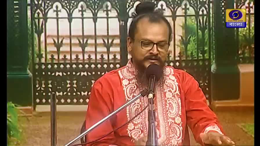 A man in a red embroidered shirt speaks into a microphone, his hands gesturing slightly. The DD Bangla logo is visible in the corner, and the backdrop suggests a studio setting.
A man in a red embroidered shirt speaks into a microphone, his hands gesturing slightly. The DD Bangla logo is visible in the corner, and the backdrop suggests a studio setting.