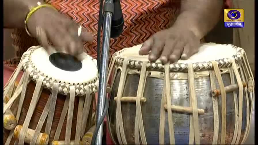 Fingers dance across the tabla drums, striking the skins with precise movements. The DD Bangla logo in the corner hints at a performance in progress.
Fingers dance across the tabla drums, striking the skins with precise movements. The DD Bangla logo in the corner hints at a performance in progress.