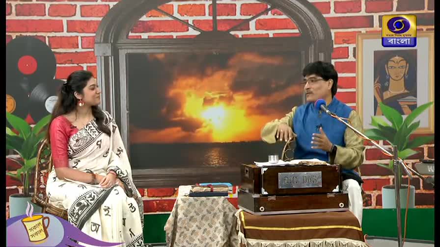 A man in a blue vest gestures with his hands, speaking into a microphone near a small harmonium. Beside him, a woman in a white sari with red sleeves listens attentively on the DD Bangla set.
A man in a blue vest gestures with his hands, speaking into a microphone near a small harmonium. Beside him, a woman in a white sari with red sleeves listens attentively on the DD Bangla set.