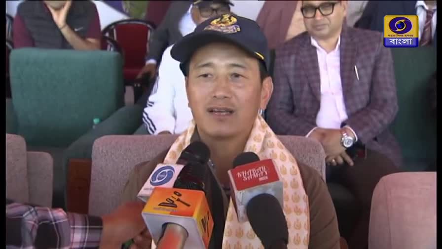 A man in a cap and scarf speaks into microphones, likely giving a press conference. The DD Bangla logo is visible in the background, suggesting this is a broadcast from India.
A man in a cap and scarf speaks into microphones, likely giving a press conference. The DD Bangla logo is visible in the background, suggesting this is a broadcast from India.