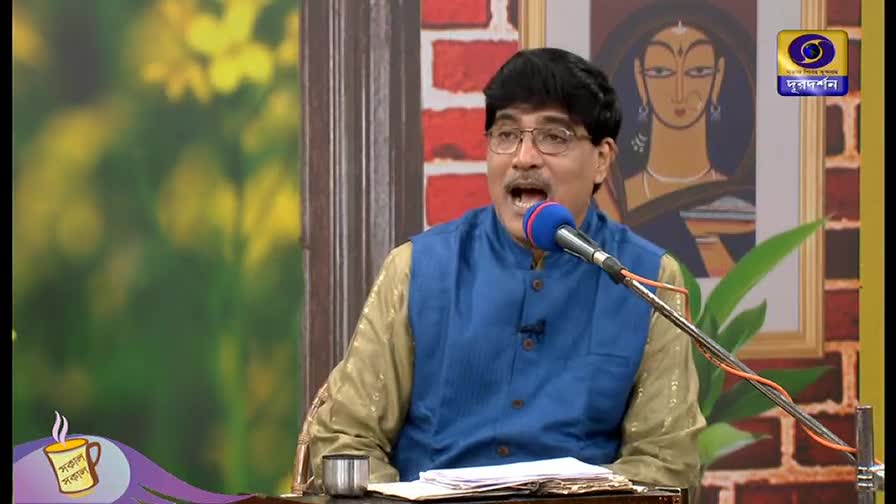 A man in a blue vest is speaking into a microphone, his mouth open. Behind him, a painting hangs on a brick wall, and the DD Bangla logo is visible in the corner.
A man in a blue vest is speaking into a microphone, his mouth open. Behind him, a painting hangs on a brick wall, and the DD Bangla logo is visible in the corner.