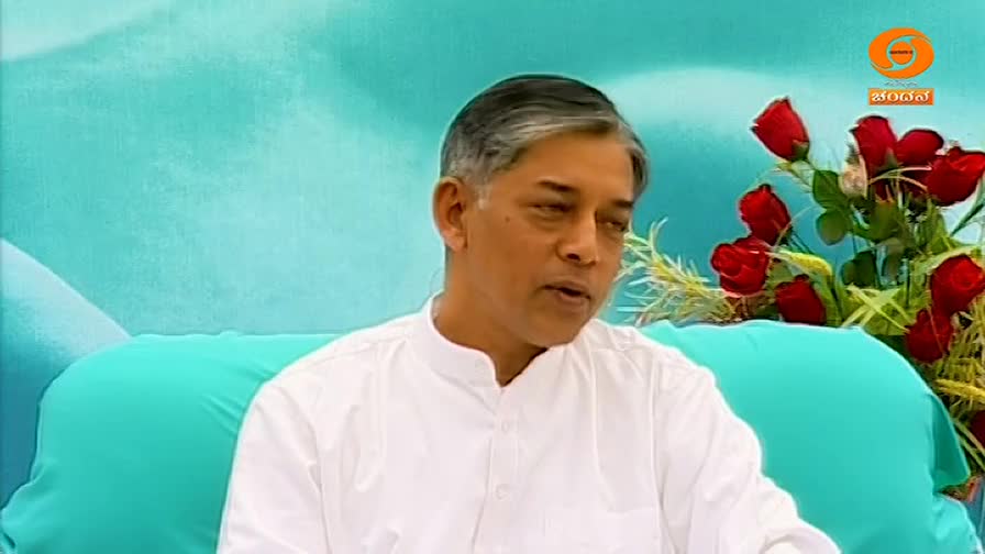 A man in a white shirt is speaking, his expression serious. Behind him, a bouquet of red roses and the DD Chandana logo add a touch of formality to the scene.
