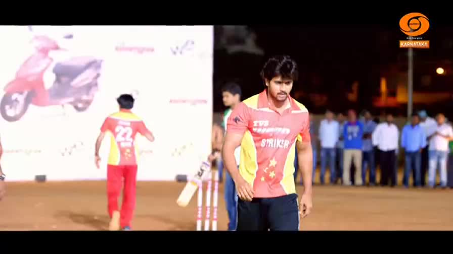 A cricketer in a red and yellow jersey stands near the wickets, looking towards the crowd. Another player, wearing the same colors, walks away from the pitch.
