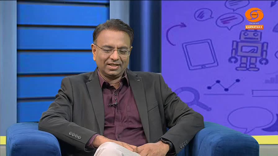 A man in a dark suit and glasses sits on a blue chair, speaking animatedly. Behind him, a purple backdrop is covered in simple drawings.
