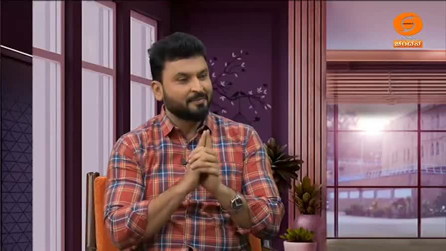 A man in a plaid shirt sits in a studio, gesturing with his hands as he speaks. The DD Chandana logo is visible in the corner, and a window with a view of the outside is behind him.
