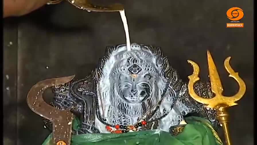 A stream of white liquid is being poured over the carved face of a deity, coating its features. The statue is adorned with jewelry and flanked by golden trident and other ceremonial objects.
