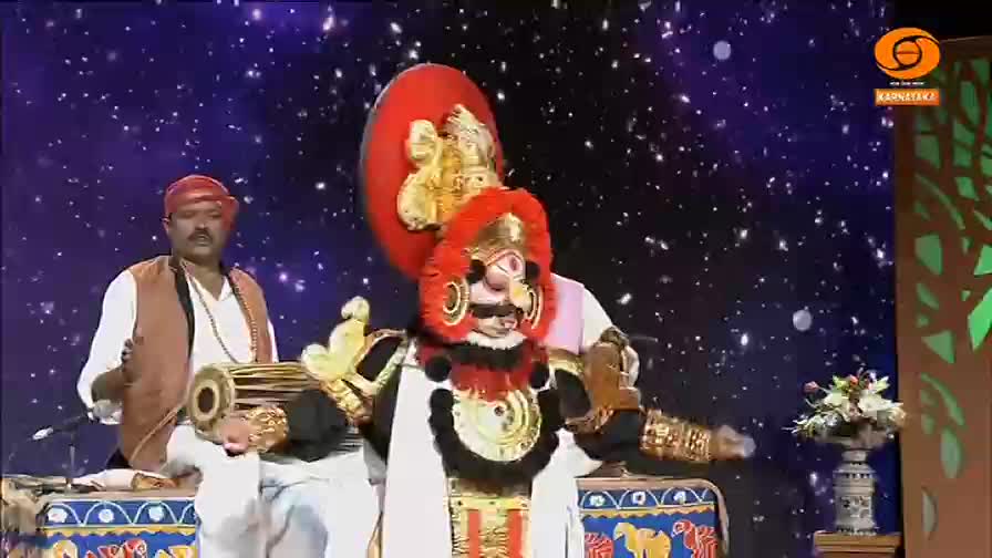 The performer, adorned in an elaborate mask and costume, is center stage, and the drummer beside him keeps time. The backdrop sparkles like a night sky on DD Chandana, a television channel in India.
