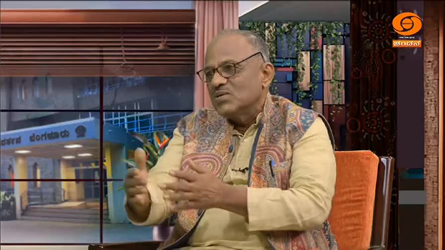 A man in glasses and a patterned vest gestures with his hands as he speaks. Behind him, a window reveals a building with a sign in Kannada, likely part of a DD Chandana broadcast in India.
