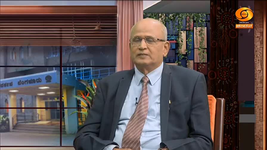 A man in a suit and tie sits in a chair, speaking on what looks like a television program. Through the window behind him, I can see a building with Kannada script.
