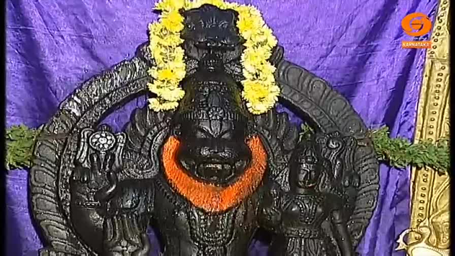 A dark statue of a deity, adorned with garlands, stands prominently against a purple backdrop. The sculpture's face is fierce, and its chest is marked with orange.
