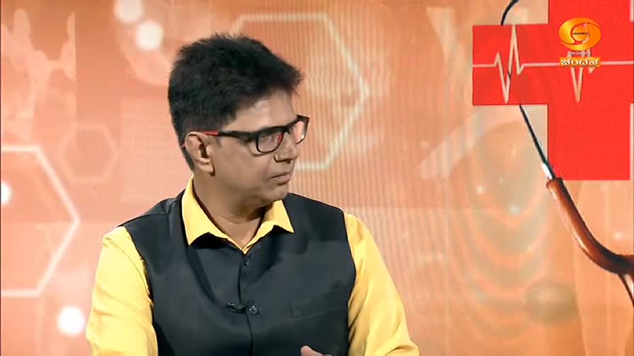 The man in the yellow shirt and black vest is looking to his left, seemingly listening intently. Behind him, a red cross and a medical symbol appear on a screen, likely from a DD Chandana broadcast in India.
