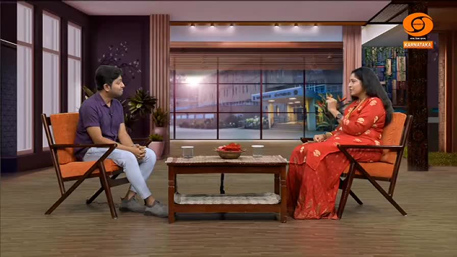A woman in a red outfit gestures as she speaks to a man in a purple shirt, both seated in chairs on a television set. The DD Chandana logo is visible in the corner, suggesting a show from India.
