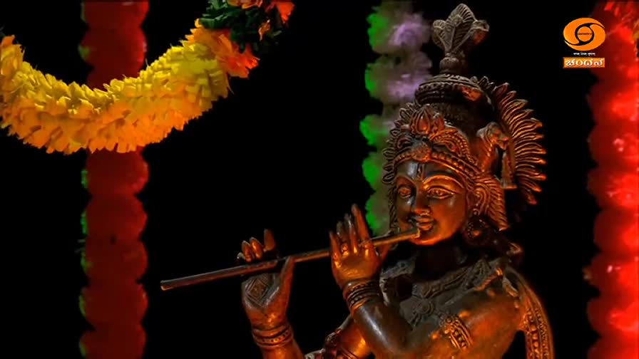 A bronze statue of Krishna plays a flute, his face illuminated against a dark background.  Garlands of yellow and red flowers hang nearby, framing the scene.
