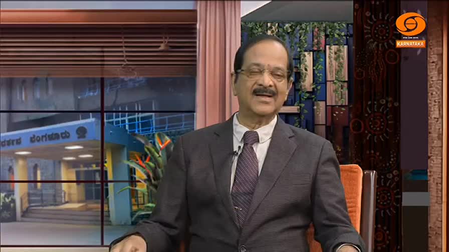 A man in a suit sits in a studio, speaking with a gentle smile. Behind him, a window shows a building, possibly a government office, in India.

