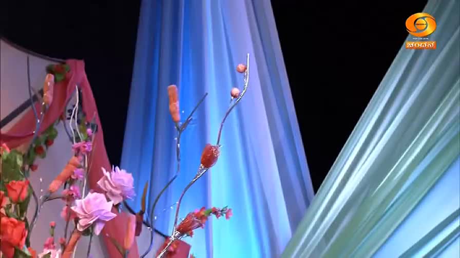 A stage is decorated with artificial flowers and fabric drapes, the backdrop a mix of pink, blue, and green. The logo for DD Chandana, a channel from India, is visible in the corner.
