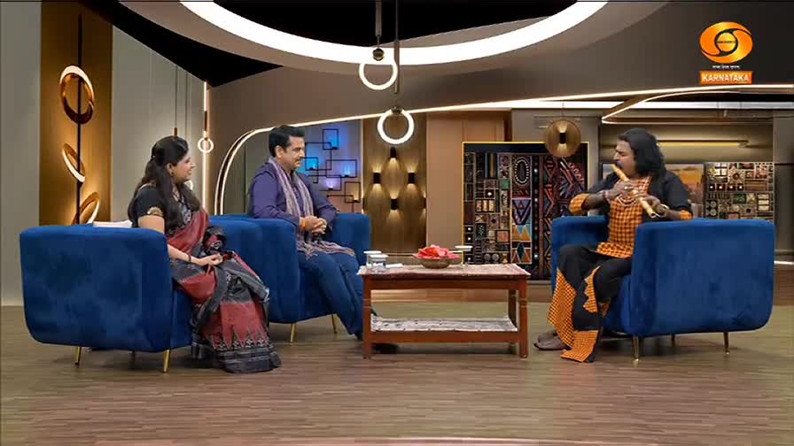 Three people sit in blue armchairs, engaged in conversation on a set for DD Chandana. A man in an orange and black patterned shirt holds a flute, presumably about to play.
