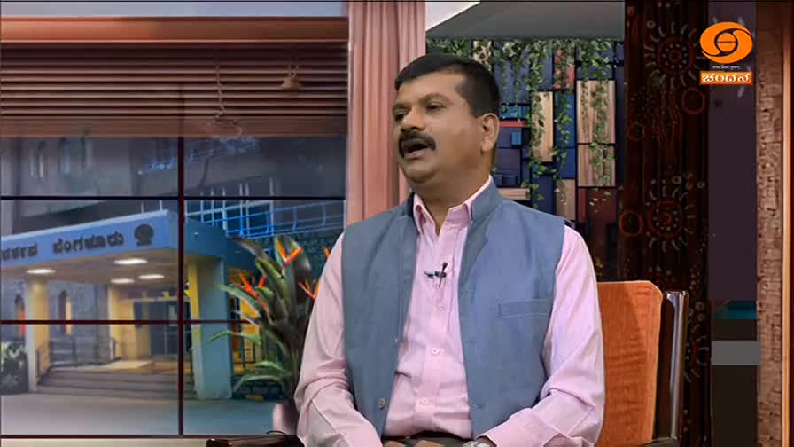 A man in a pink shirt and vest is speaking, the DD Chandana logo visible in the corner. Behind him, a window shows a building and some greenery.
