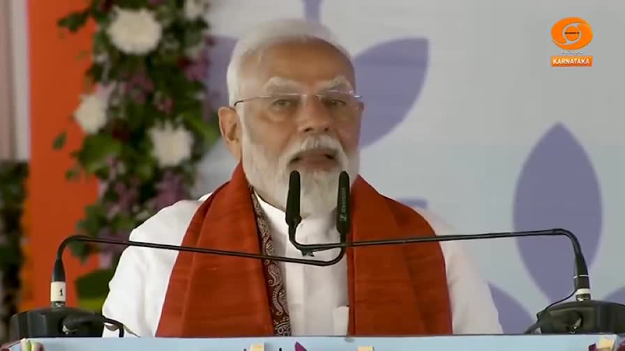 A man with a white beard and glasses speaks into two microphones, his mouth open as he addresses the crowd. He wears a white shirt and an orange scarf, the backdrop a muted blue and white.
