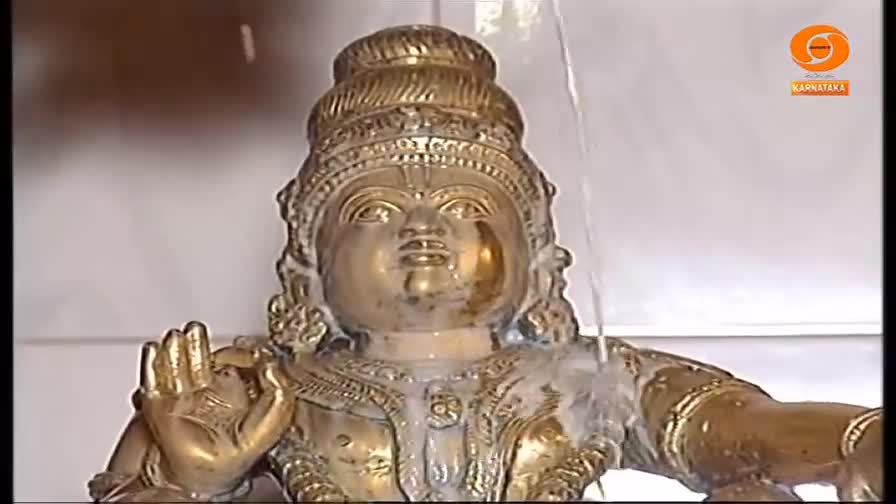 Water streams down the surface of a golden statue, washing over the figure's face and arms. The scene is likely from a religious ceremony, possibly broadcast on DD Chandana in India.
