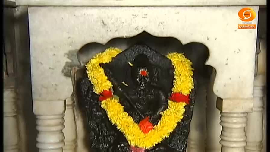 A dark statue, adorned with a vibrant yellow garland, stands inside a white-pillared structure. The scene appears to be part of a broadcast from DD Chandana in India.

