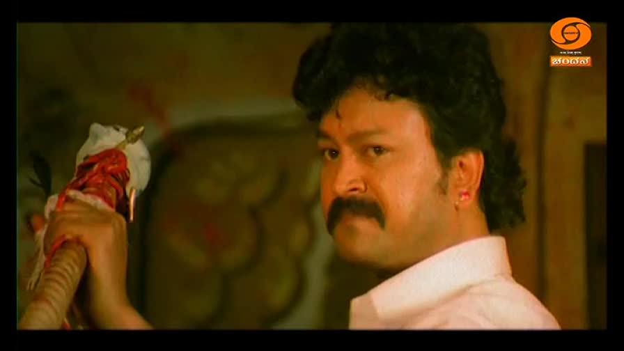 A man with a thick mustache stares intently, holding a staff with a bloody, white object impaled on top. The DD Chandana logo is visible in the corner, and the scene has a dramatic, slightly unsettling feel.
