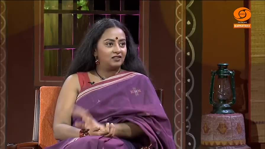 A woman in a purple saree, likely DD Chandana, sits in a chair, gesturing with her hands as she speaks. Behind her, a decorative wall and a lantern add to the studio's ambiance.

