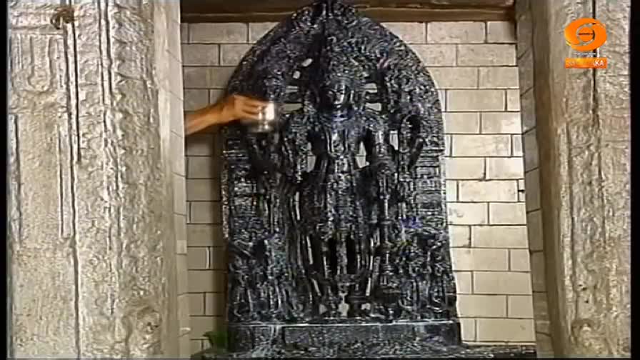 A hand pours liquid from a small metal vessel onto a dark, intricately carved statue of a deity. The statue stands inside a stone alcove, its details highlighted by the liquid.
