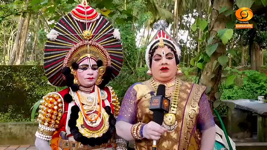 Two performers in elaborate costumes stand side-by-side, one holding a microphone with the DD Chandana logo. The person on the right is speaking, their face heavily made up, while the other looks on, their headpiece a striking fan of red, black, and gold.
