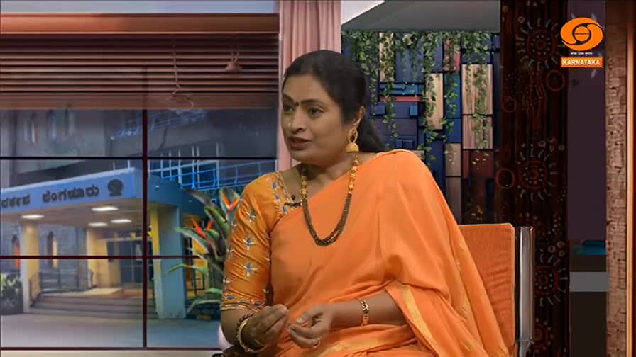 A woman in an orange sari sits in a studio chair, speaking animatedly. The background shows a window with a building visible outside and a logo for DD Chandana.
