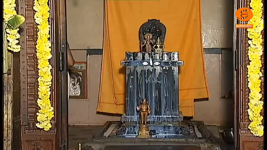 A small golden statue stands on a pedestal, with a larger, dark stone structure behind it. Yellow garlands frame the scene, and an orange cloth hangs behind the central figures.
