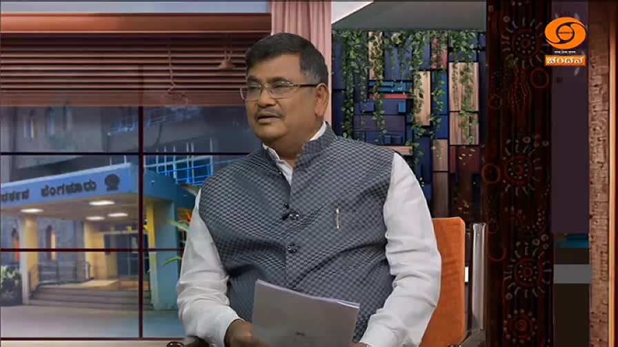A man in a patterned vest sits in a studio, holding a paper and speaking. Behind him, a window shows a building and the DD Chandana logo is visible on the right.
