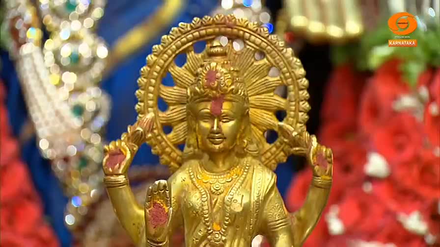 A golden idol, adorned with a halo, is the focal point, its hands raised in a gesture of blessing. The background is a mix of soft focus colors, likely from a television broadcast on DD Chandana in India.
