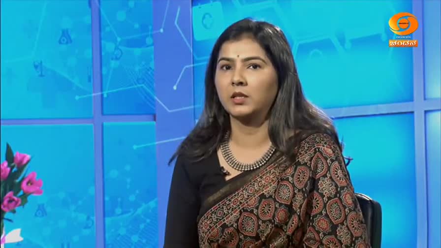 A woman in a patterned saree sits in a studio, speaking directly to the camera. Behind her, a blue backdrop with a network design is visible.
