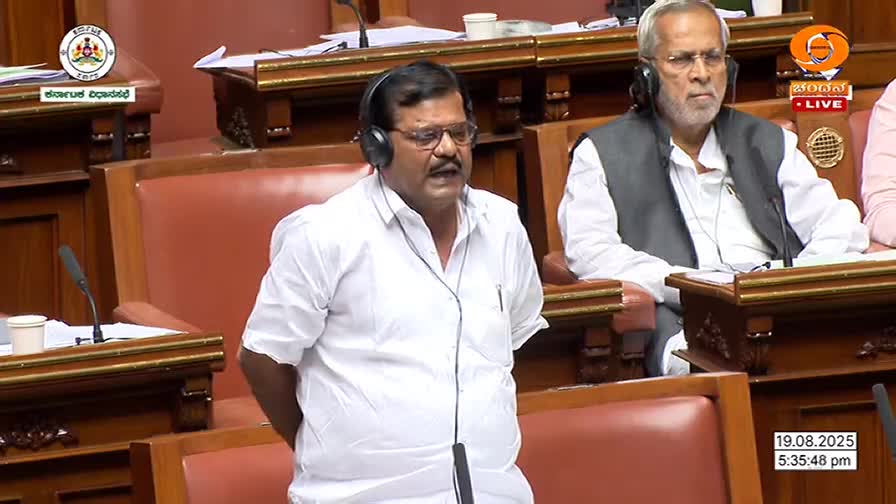 A man in a white shirt and glasses speaks, his mouth open as if mid-sentence.  He wears a headset, and the DD Chandana logo is visible in the corner, indicating a live broadcast from India.
