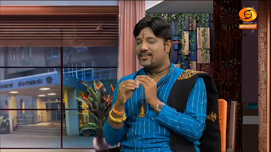 A man in a blue shirt and gold jewelry gestures with his hands while speaking. Behind him, a window offers a glimpse of a building and some greenery.
