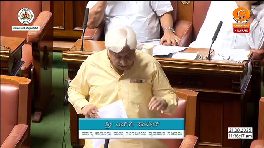 A man in a yellow shirt, with white hair, is speaking from a wooden desk in a chamber. Papers are in his hands as he gestures, likely addressing the room.
